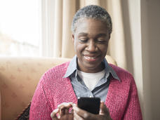 Woman with short hair sitting comfortably in a chair by a window, smiling while using a smartphone. She is wearing a pink cardigan over a denim shirt. 