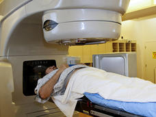 A patient in a hospital gown lies on a treatment table beneath a large linear accelerator machine during a radiation therapy session.