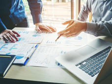 A close up of two peoples hands on a desk that is covered with financial charts. One of the hands is pointing towards one of the chart pages. An open laptop is in the foreground.
