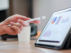 Close up hand with a stylus pen pointing on tablet screen that shows a pie chart, table and bar graphics in a conference room setting