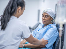 A nurse holds the arm of a sitting patient: an elderly woman with a head covering. A blurry IV bag is in the foreground; the background indicates a treatment center.