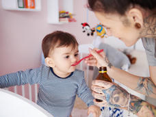 A smiling tattooed woman gives a child a spoonful of dabrafenib. The child is standing in a crib and the background indicates a child’s bedroom.