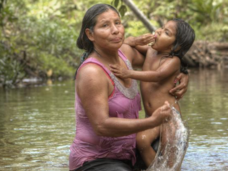 A woman in a sleeveless top and pants carries a child, who is eating, through a river. Both the woman and the child are soaked. The background is filled with green plants and trees, indicating a forest or jungle.