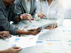 Four individuals gathered around a table in an office setting discussing data displayed on a tablet, surrounded by printed charts and graphs. 