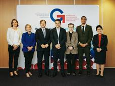 Group of seven individuals stand side by side in front of a backdrop featuring the G7 Cancer logo  and the slogan "Accelerate the Progress Against Cancer Worldwide." They are dressed in formal business attire, including suits and blazers. Behind the backdrop is a wooden wall background.