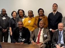 A group of people in professional clothing with name badges look at the camera and smile. The majority of the people in the Photo are black; there are two white men and one Asian woman. Three men are seated, and the rest of the group is standing. 