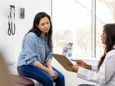 An adult female patient sits on an exam table in a clinic exam room, looking at a medical chart held by a medical provider.