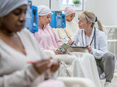 Three women sit in a clinic having IV infusions. A health provider kneels beside and chats with one young woman who is looking at a pamphlet.
