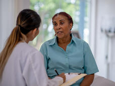 A black woman speaks to a younger woman wearing a white doctor's coat and holding a clipboard and pen.