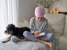 Una niña con un pañuelo rosado en la cabeza escribe en un cuaderno con un perro a su lado.