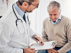 An older man in a v-neck sweater and button down shirt, seated, speaking with a doctor with glasses and a beard.