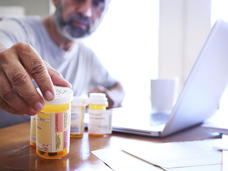 Photo of a man reaching for prescription pill bottles sitting on a table in the foreground.