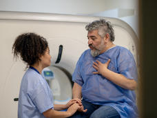 An older man with a beard, wearing a medical gown, hand on his chest, speaking to a woman radiology technician in scrubs