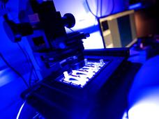 A photograph of a square-shaped device with small channels, illuminated in white light with a large microscope hanging over top, that can measure the stickiness of tumor cells.