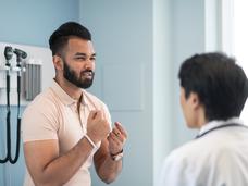 A young man with dark black hair and beard, form fitting orange shirt, with his hands in front of him speaking with a doctor. 