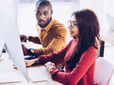 A man wearing a yellow sweater working alongside a woman wearing glasses and a red sweater. They are in front of a large screen computer and the man is pointing to the screen.