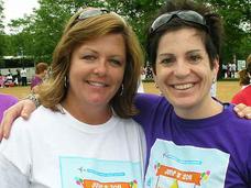 Two women stand looking at the camera and smile. Both are wearing sunglasses on top of their heads and have different colored shirts with the same graphic treatment on them. The woman on the left has shoulder length light brown hair and is wearing a white shirt; the woman on the right has short black hair and is wearing a purple shirt.