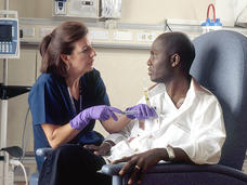 A nurse administers chemotherapy to a patient. Both are seated. The nurse has shoulder-length brown hair and wears dark teal scrubs and purple gloves. She is holding a syringe that is held against the patients chest. The patient is wearing casual clothing and he looks at the nurse.
