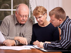 An elderly couple look down at papers on a table with a younger man. The elderly man is bald with a mustache and wears glasses with a button up shirt. The woman has short dirty blonde hair with glasses and a dark top. The younger man has short light brown hair and a striped button up shirt.