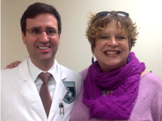 Dr. Christian Hinrichs and Susan Scott stand looking at the camera and smile. Dr. Hinrichs has short dark hair, is wearing glasses, a white coat, and shirt and tie. Susan has short, dirty blonde hair, is wearing sunglasses on top of her head and a large purple scarf and top.