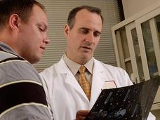 A male doctor holds an x-ray he is looking at while speaking to a male patient. The doctor has short dark hair and wears a white lab coat with shirt and tie. The patient has a yellow-and-black polo shirt. Window blinds and a cabinet are in the background.