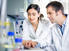 Two scientists wearing lab coats are collaborating at a computer in a laboratory setting