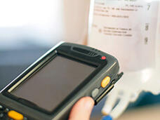 Close-up view of a handheld electronic scanning device with a small screen and yellow buttons being used to scan a barcode or QR code on a medical label.