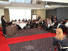 A group of people sitting at tables in a conference room watching a presenter.