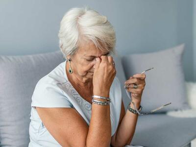 An elderly woman sits on a couch holding her glasses in one hand and rubbing her forehead with the other, appearing to be in discomfort.