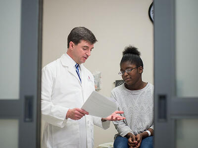 A doctor looks at a piece of paper and speaks to his patient. The doctor is a white middle-aged male, wearing a lab coat, button up shirt and tie, He is standing and gesturing as he speaks. The patient, a younger Black woman with glasses, a long sleeve grey sweater and jeans, sits on the exam table and looks slightly distressed. Out of focus doors to the room are in the foreground.