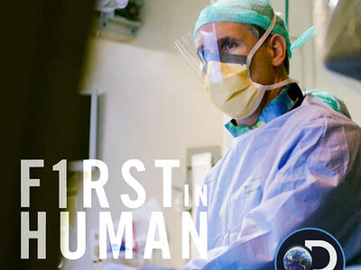 A doctor in an operating room wears a plastic face shield, a mask, scrubs and a green cap. He looks off into the distance. In the bottom left corner, text reads “F1RST in HUMAN” with the dates and times of August 10, 17 & 24, 9pm ET/PT. The Discovery Channel logo is on the bottom right corner.