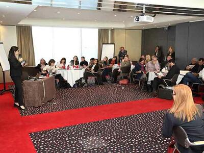 A group of people sitting at tables in a conference room watching a presenter.