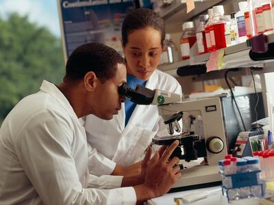 A male researcher looking through a microscope in a laboratory with a female researcher standing near.