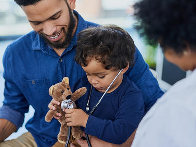 A pediatric patient wears a stethoscope and holds a stuffed bear as his father smiles at him