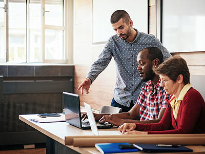 Two adults work on laptops while another offers assistance