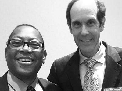 Black and white photo of two men in suits standing side-by-side and smiling at the camera. 