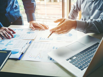 A close up of two peoples hands on a desk that is covered with financial charts. One of the hands is pointing towards one of the chart pages. An open laptop is in the foreground.