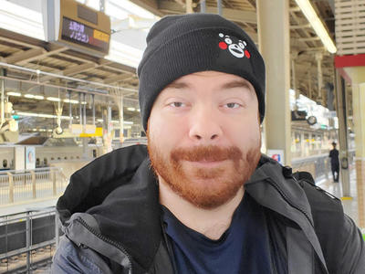 Headshot of a light-skinned man, Justin, in a black hat, black and grey jacket, and blue shirt