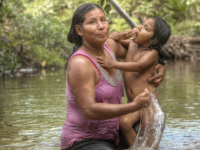 A woman in a sleeveless top and pants carries a child, who is eating, through a river. Both the woman and the child are soaked. The background is filled with green plants and trees, indicating a forest or jungle.