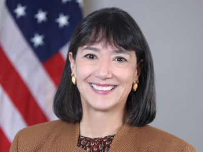 Woman (NCI Director Dr. Monica Bertagnolli) with dark hair wears a brown jacket over a patterned top and smiles at the camera.