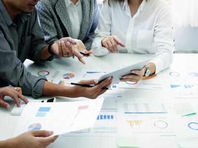 Four individuals gathered around a table in an office setting discussing data displayed on a tablet, surrounded by printed charts and graphs. 