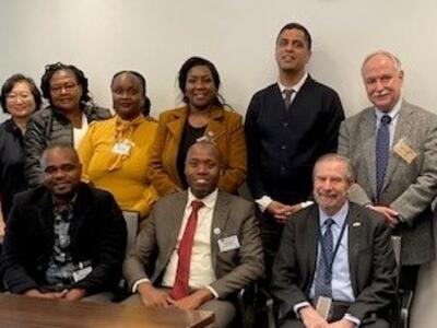 A group of people in professional clothing with name badges look at the camera and smile. The majority of the people in the Photo are black; there are two white men and one Asian woman. Three men are seated, and the rest of the group is standing. 