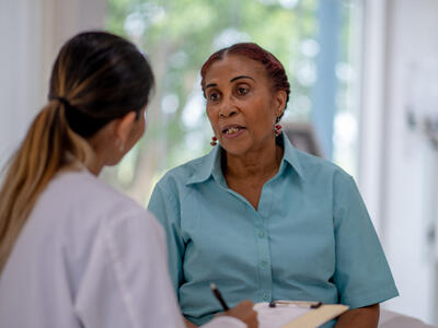 A black woman speaks to a younger woman wearing a white doctor's coat and holding a clipboard and pen.