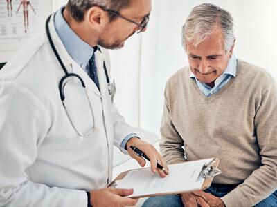 An older man in a v-neck sweater and button down shirt, seated, speaking with a doctor with glasses and a beard.