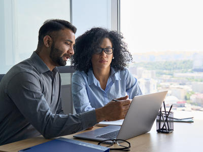 Indian male ceo executive manager mentor giving consultation on financial operations to female African American colleague intern using laptop sitting in modern office near panoramic window.