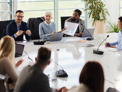 People in a meeting room at a large white table, one holding a chart. Laptops and papers are on the table; a large window is in the background.