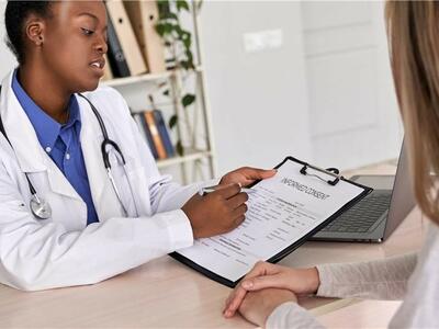 Clinician and patient sitting across from each other at a desk. The clinician is holding a clipboard with a paper that reads Informed Content at the top. The patient has her hands folded together as she listens to the clinician explain the document and signature requirements.