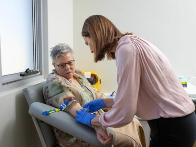Health care provider wearing blue gloves performs a blood draw on a seated patient in a clinic exam room.