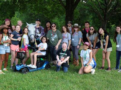 A large group of Camp Fantastic participants poses outdoors with a white horse under leafy trees, with one person seated on a mobility scooter.