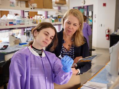 A laboratory researcher examines a sample while a colleague observes and takes notes.